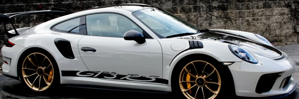 A sleek white sports car with "GT3 RS" on its side, featuring gold rims and a black spoiler, parked against a stone wall. The scene exudes speed and luxury.