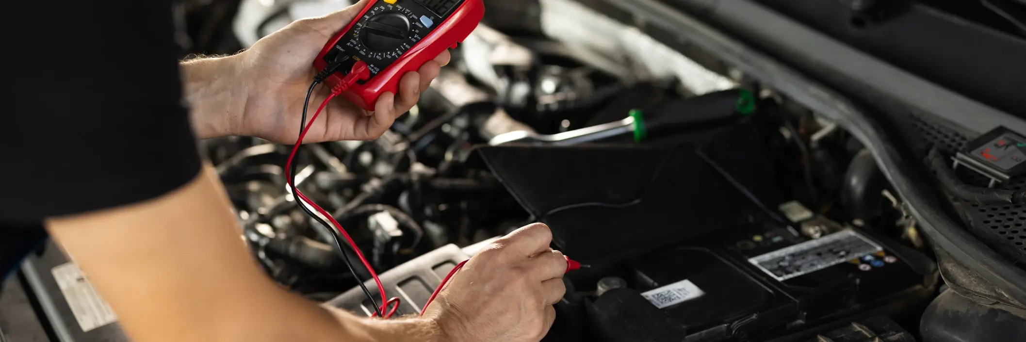 A person uses a red multimeter to check a car battery under the hood. The scene conveys a sense of focus and technical precision in automotive maintenance.