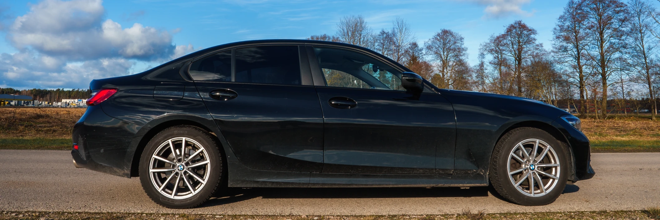 A sleek black sedan is parked on a rural road, surrounded by bare trees under a partly cloudy blue sky, conveying a serene and peaceful atmosphere.