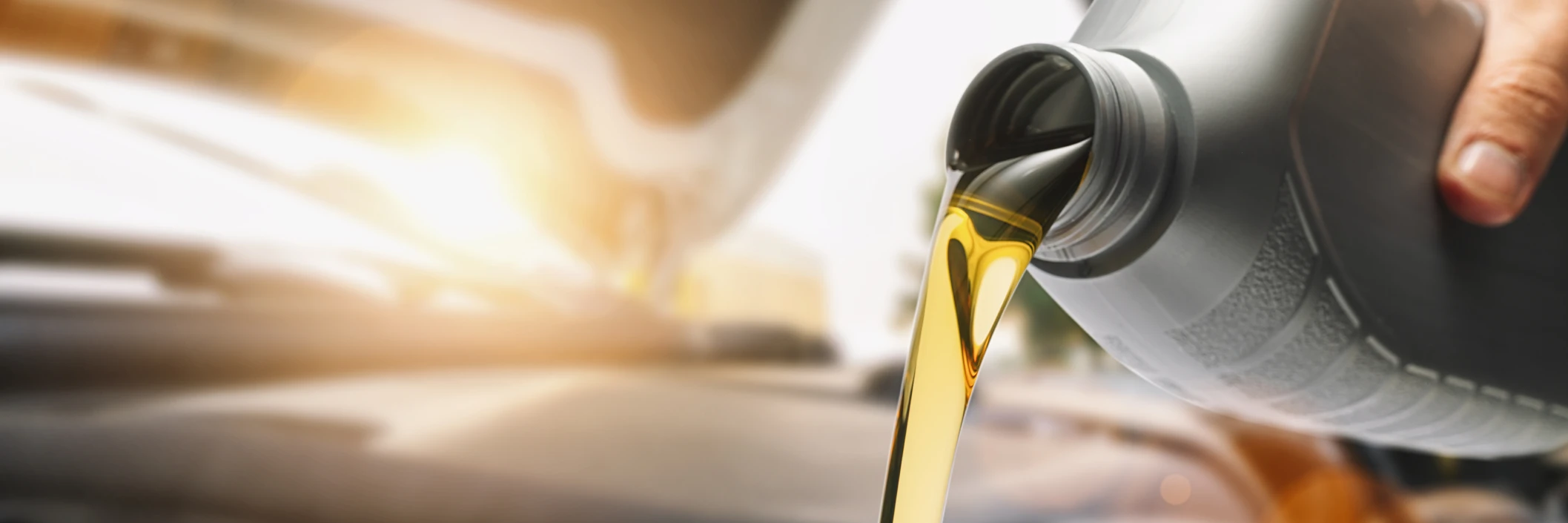 Hand pouring motor oil from a container into a car engine with the hood open. Sunlight shines brightly, conveying a clean and efficient maintenance scene.