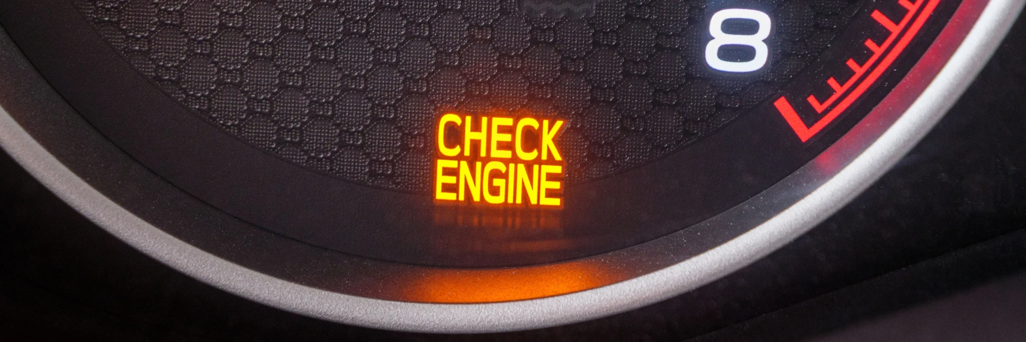 Close-up of a car dashboard showing a lit "Check Engine" warning light in bright orange. The gauge displays a number 8 and a partial red scale.