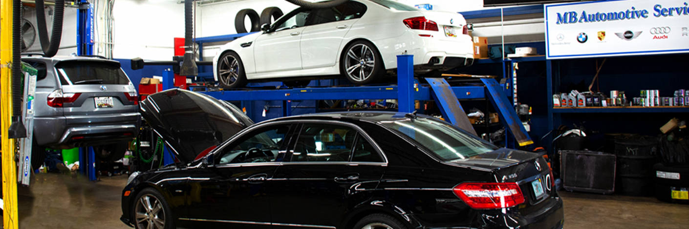 Dealership vs. Independent European Auto Repair Shop: Which is Better for Your Vehicle in Rockville, Potomac, Bethesda, North Bethesda, MD. with MB Automotive Services. Image of Black Mercedes car with hood openn, Gray BMW SUV and White BMW Sedan on car lifts in shop with tools and shop service signage in the background.