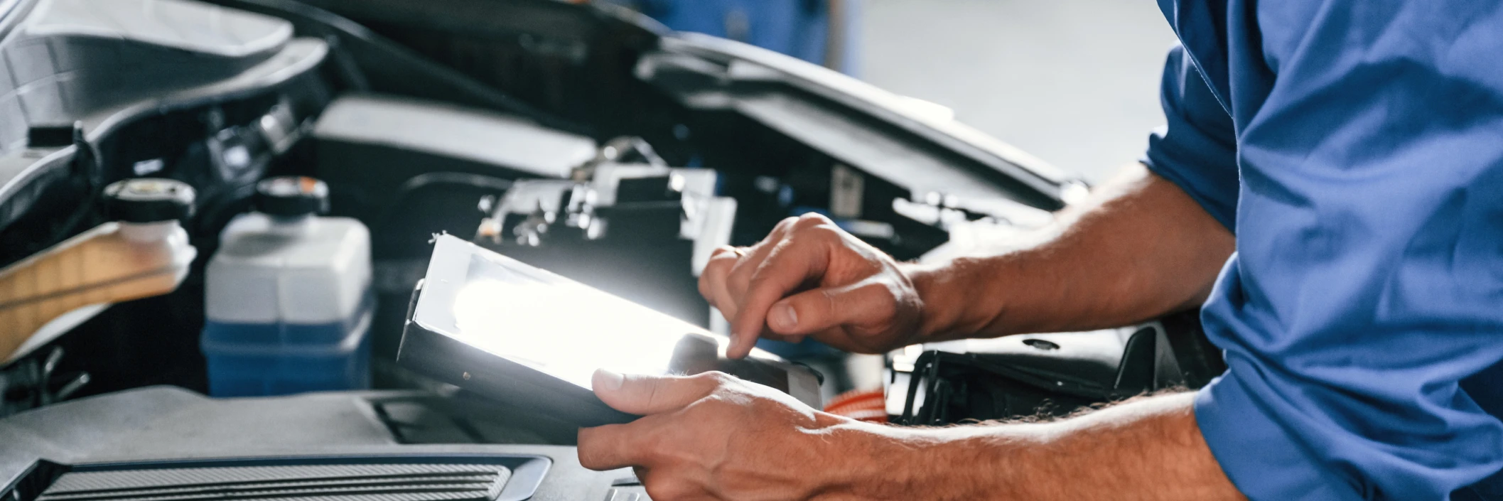 A mechanic in a blue shirt uses a tablet to diagnose a car engine. The setting conveys a modern, professional, and focused atmosphere.