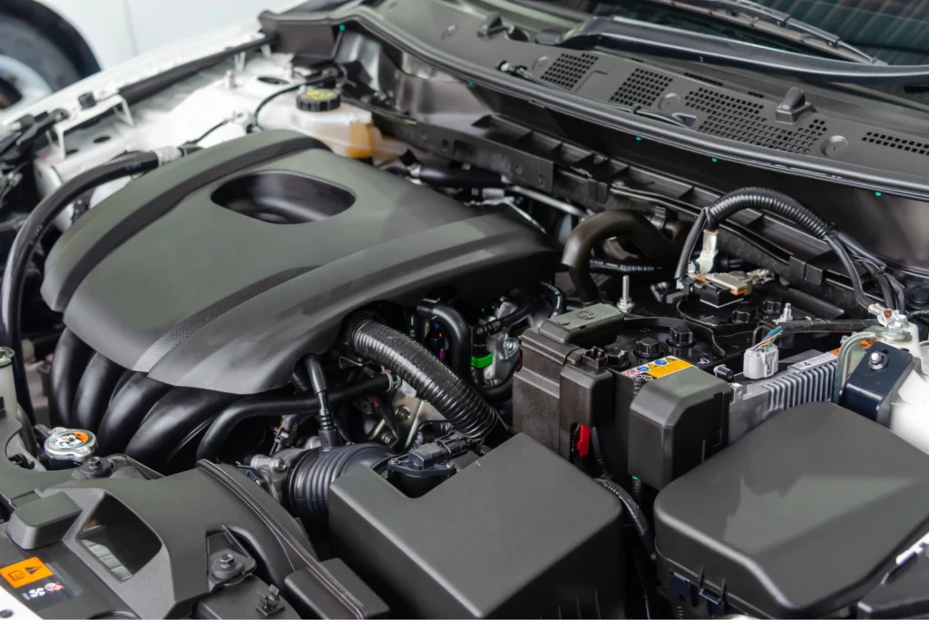 Close-up of a car engine bay, featuring a sleek black engine cover. Visible components include cables, hoses, and a battery in a clean setup.