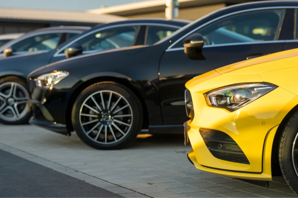 A row of sleek, modern cars parked on a sunlit, tiled lot. Foreground shows a striking yellow car, with shiny black vehicles behind, conveying luxury and style.