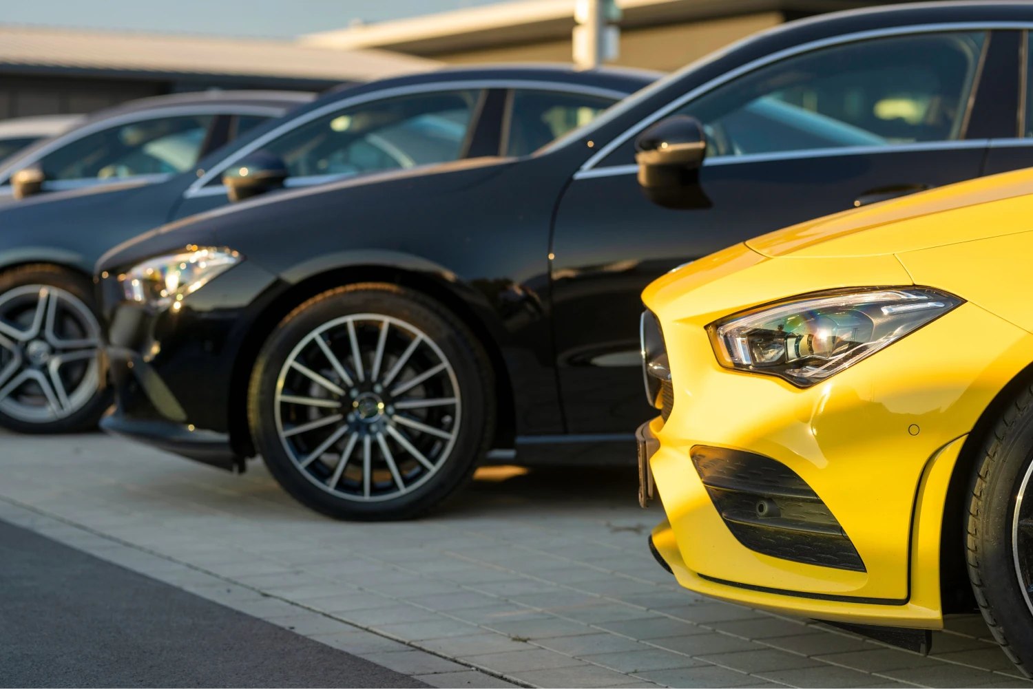 A row of sleek, modern cars parked on a sunlit, tiled lot. Foreground shows a striking yellow car, with shiny black vehicles behind, conveying luxury and style.